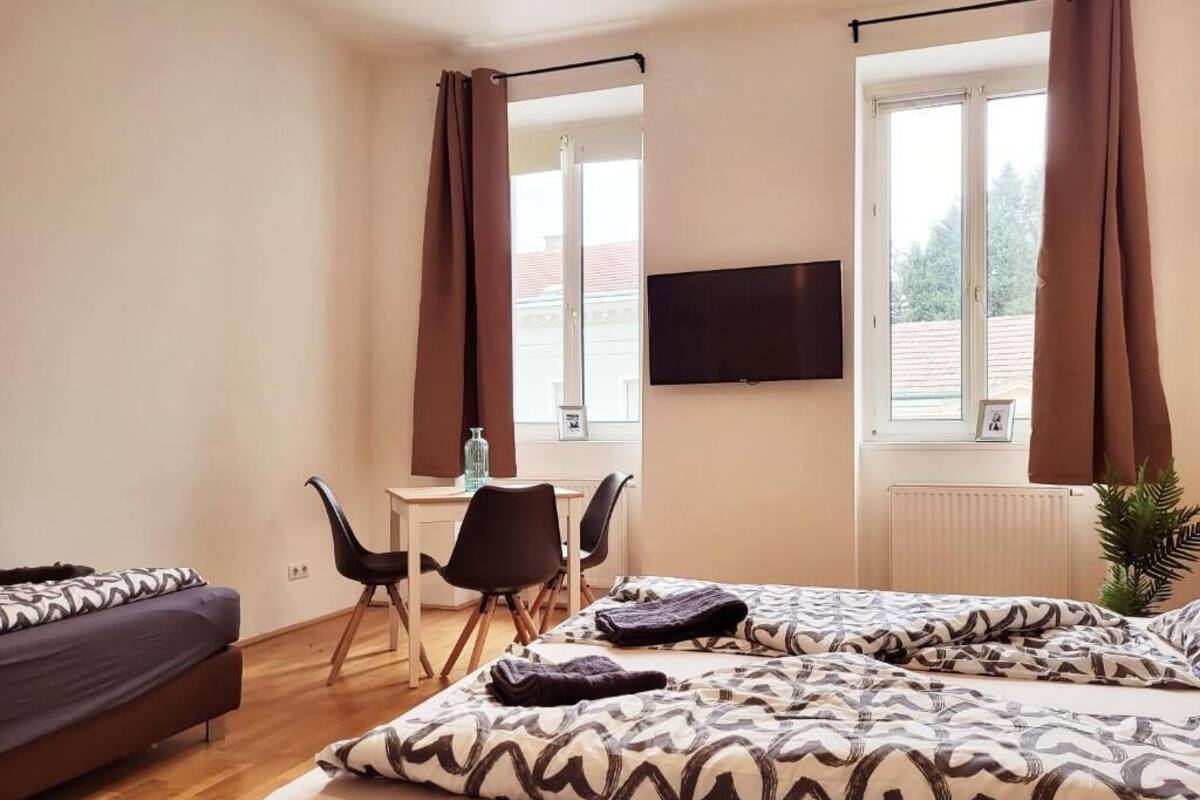 A hotel room with a bed, a desk, two chairs, a TV, and large windows with curtains.