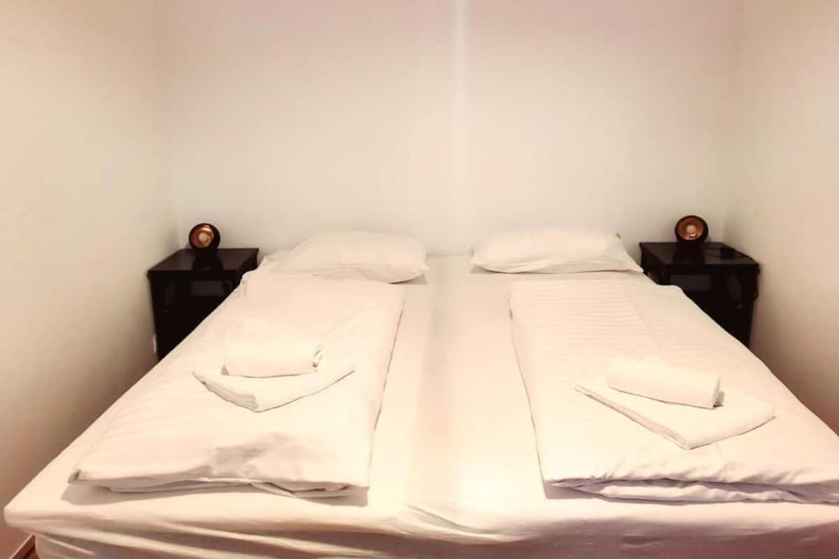 A double bed with white linens, two bedside tables with clocks, and a plain wall.