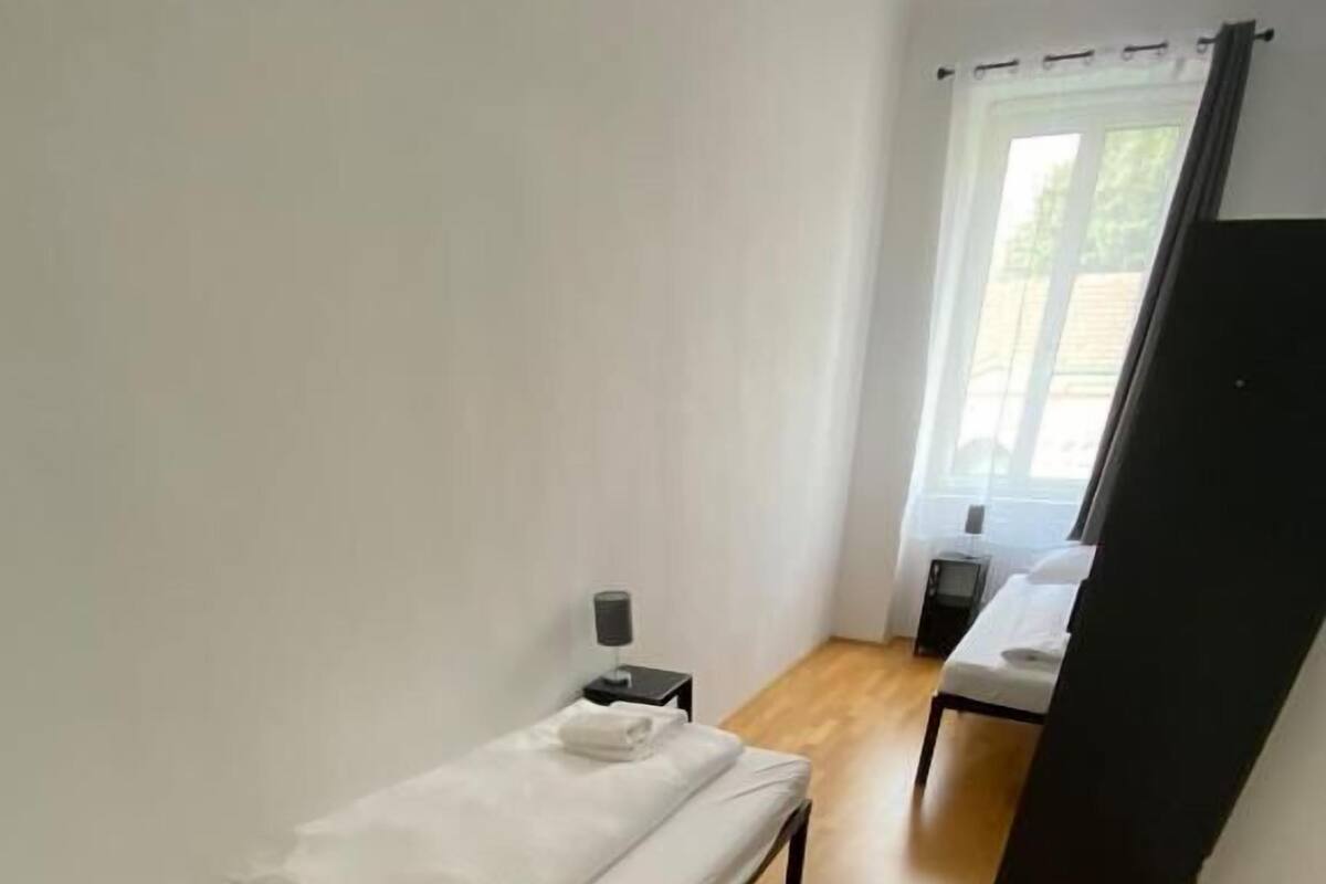 A small, single-bed room with a wooden floor, a nightstand with a lamp, and a window with curtains.