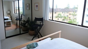 A bedroom with a bed, a desk, a chair, and a large window with a view of buildings and trees. - Tranquil, Stylish, Retreat - 3 Bedrooms (Tuncurry)