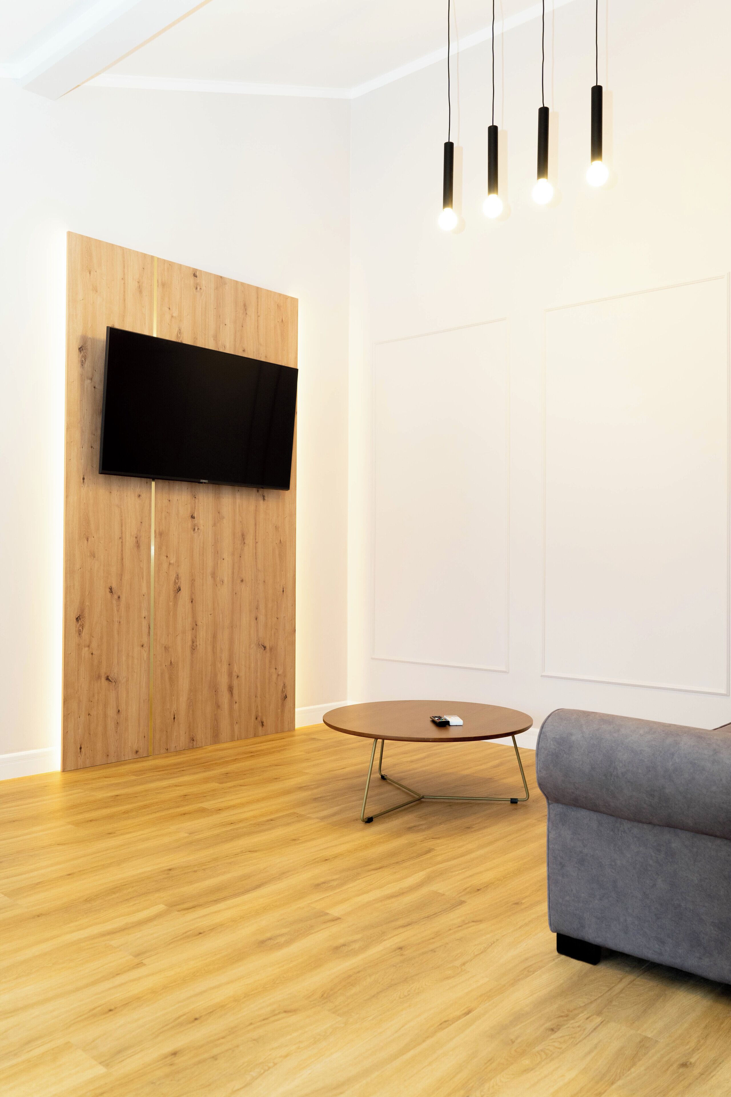 A modern living room with a flat-screen TV mounted on a wooden panel wall, a round wooden coffee table, and a gray sofa.
