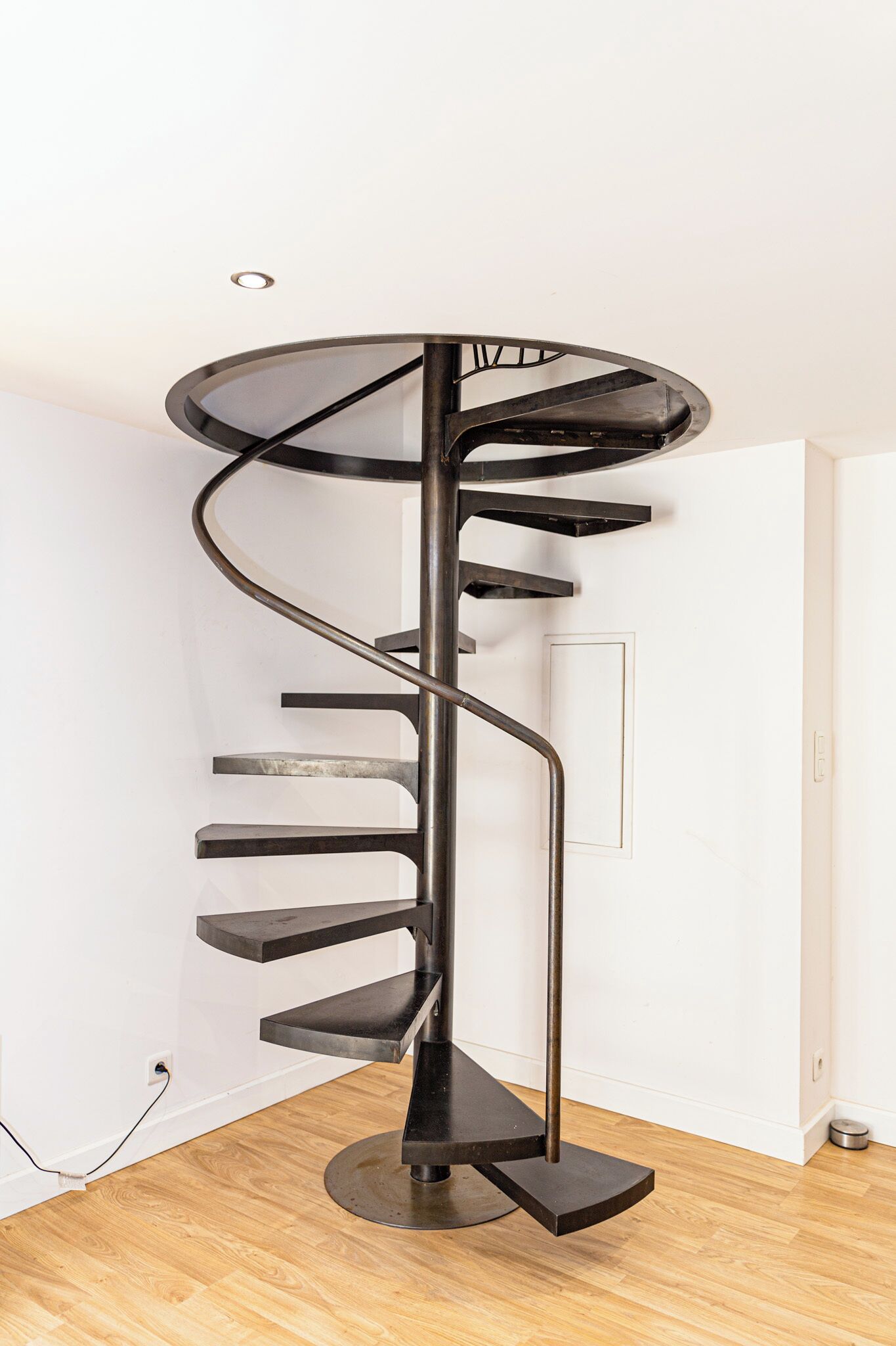 Un escalier en colimaçon au design moderne, fait de métal et de verre, dans une pièce dotée d’un plancher en bois et de murs blancs.