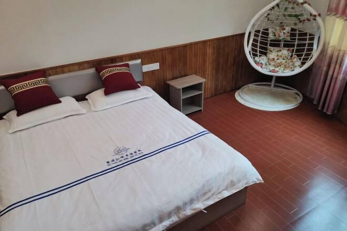 A bed with white bedding and red pillows, a wooden headboard, a small bedside table, and a hanging chair with a floral pattern.