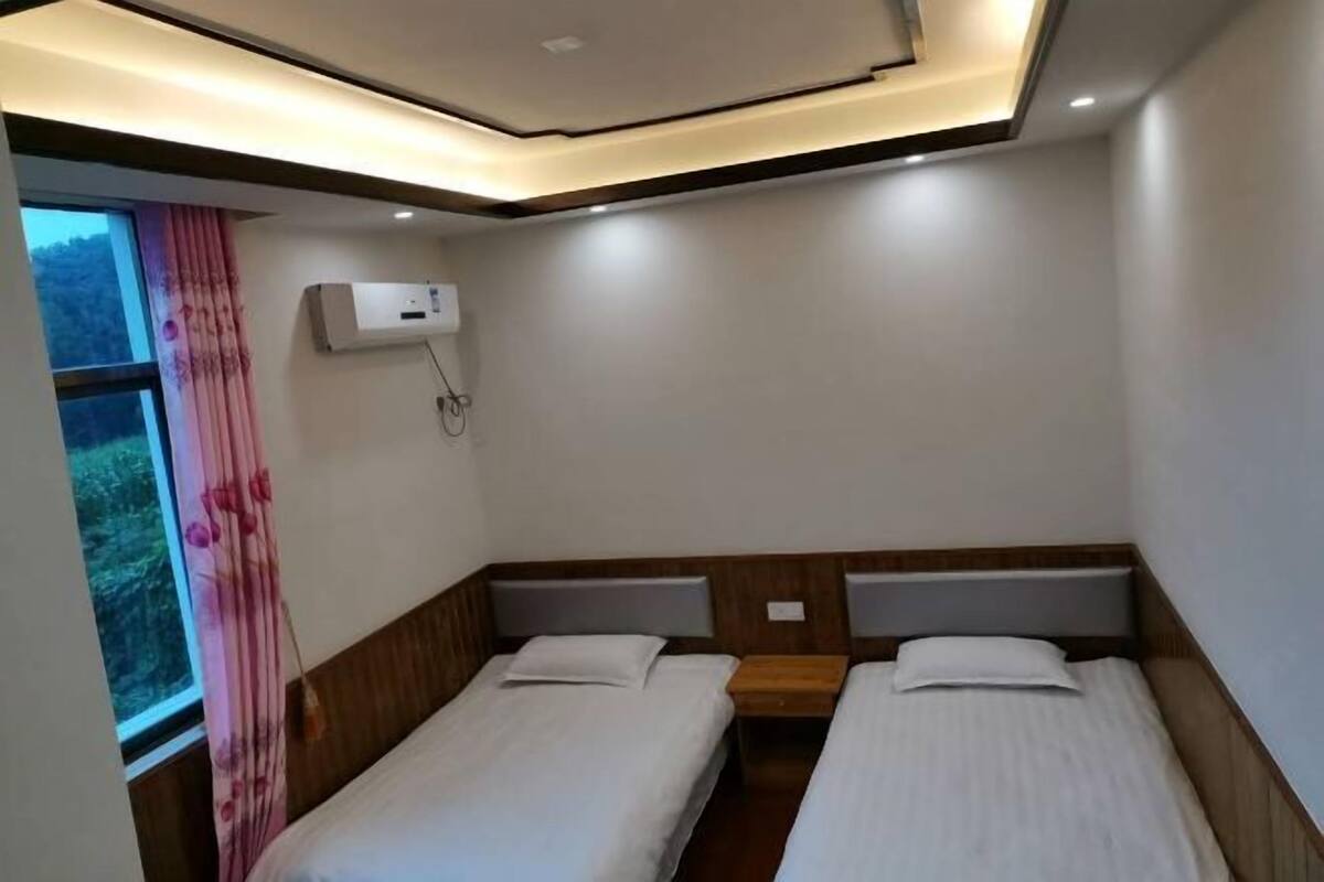 A hotel room with two single beds, a window with curtains, an air conditioning unit, and a wooden headboard.