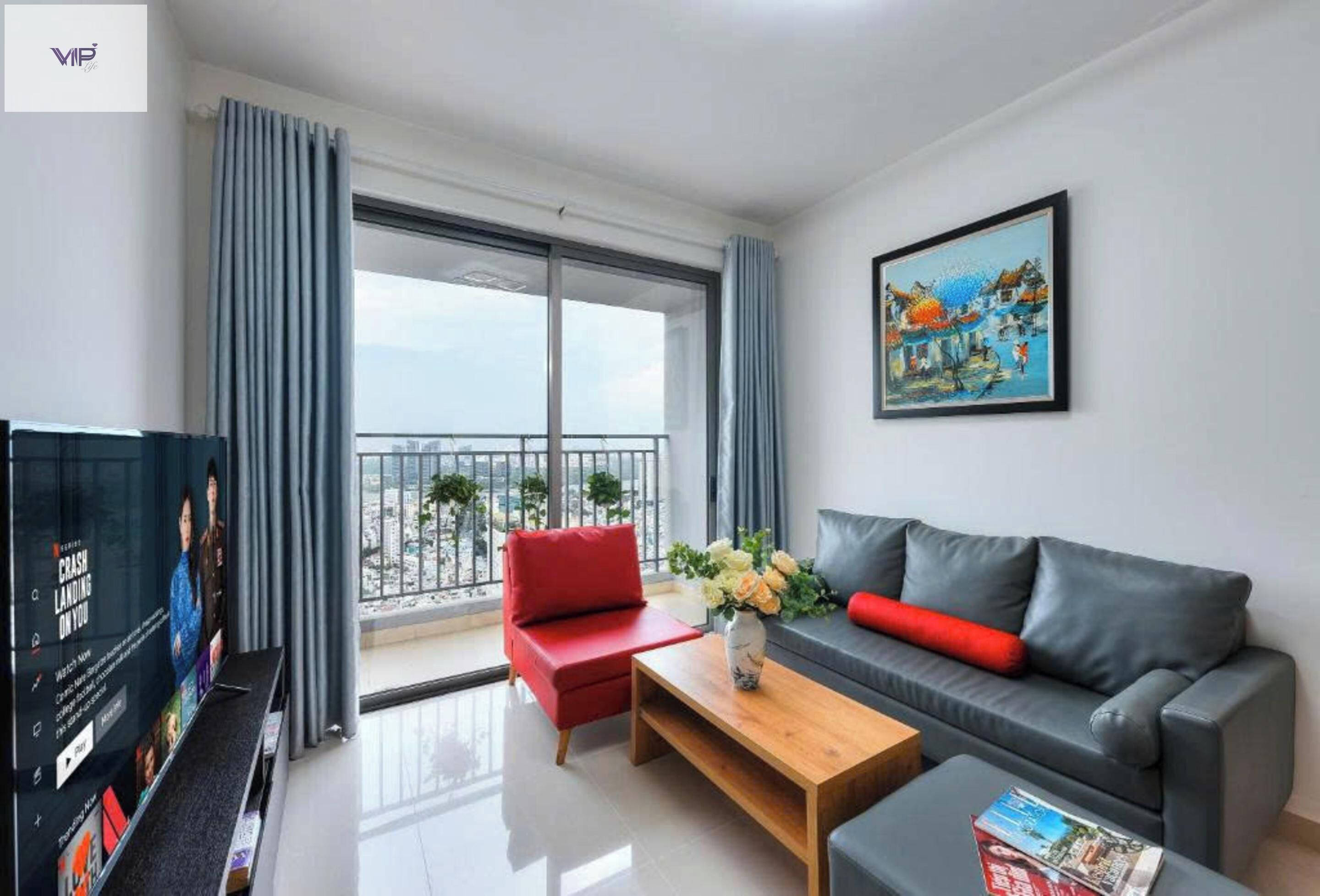Una sala de estar moderna con un sofá gris, un sillón rojo, una mesa de centro de madera y un televisor a la izquierda. Se ve un balcón a través de un ventanal grande.