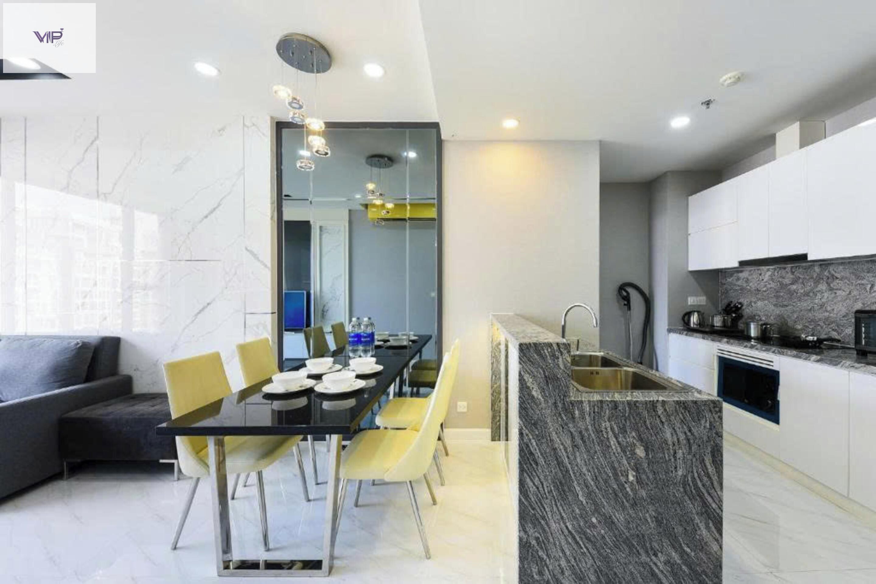 A modern kitchen with a marble countertop, a dining area with a glass table and yellow chairs, and a grey sofa in the living area.