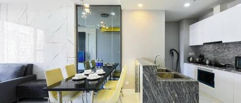 A modern kitchen with a marble countertop, a dining area with a glass table and yellow chairs, and a grey sofa in the living area.