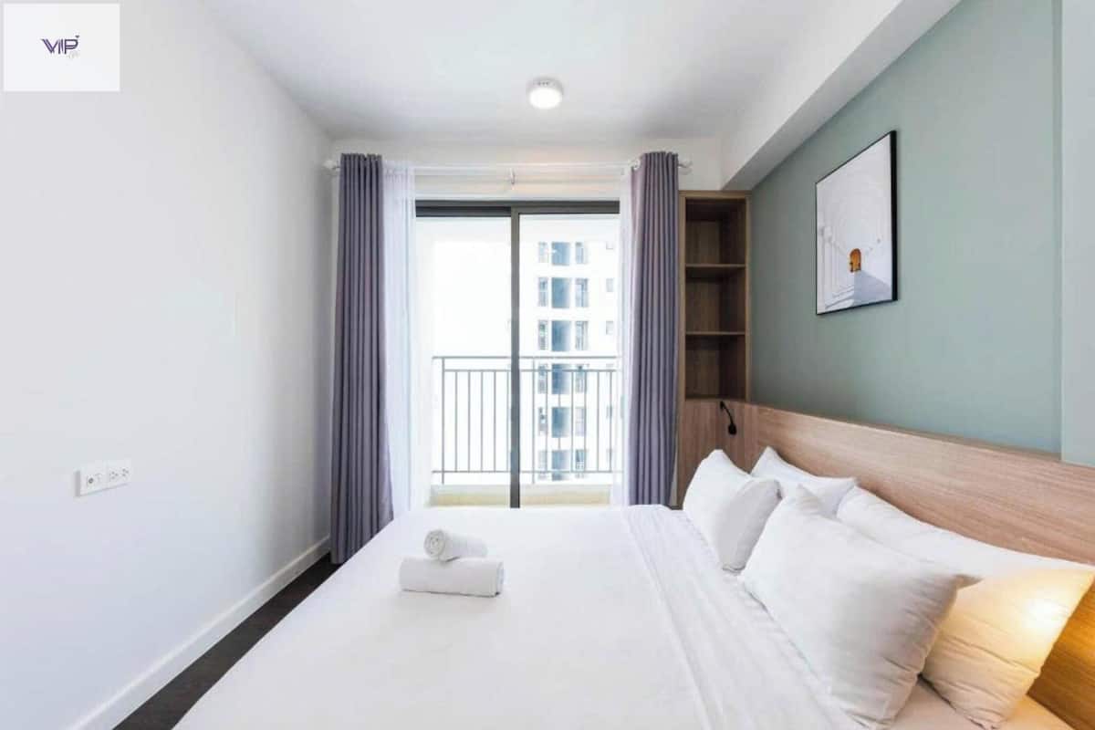 A hotel room with a large bed, a headboard, a window with curtains, a framed picture on the wall, and a view of a balcony and building outside.