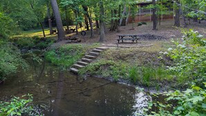 Een bebost gebied met een picknicktafel, een houten bruggetje over een beekje, en een gebouw op de achtergrond.