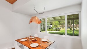 Un coin repas avec une table en bois dressée pour quatre personnes, des chaises blanches et un luminaire suspendu. Une grande fenêtre offre une vue sur un quartier résidentiel avec des arbres et des maisons.