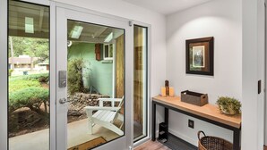Un intérieur moderne avec une porte-fenêtre coulissante donnant sur un jardin, une chaise Adirondack blanche, un banc en bois, une petite table avec des bougies et un tableau encadré au mur.