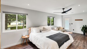 Une chambre moderne avec un grand lit, deux fenêtres, un ventilateur de plafond et une vue sur l’extérieur.