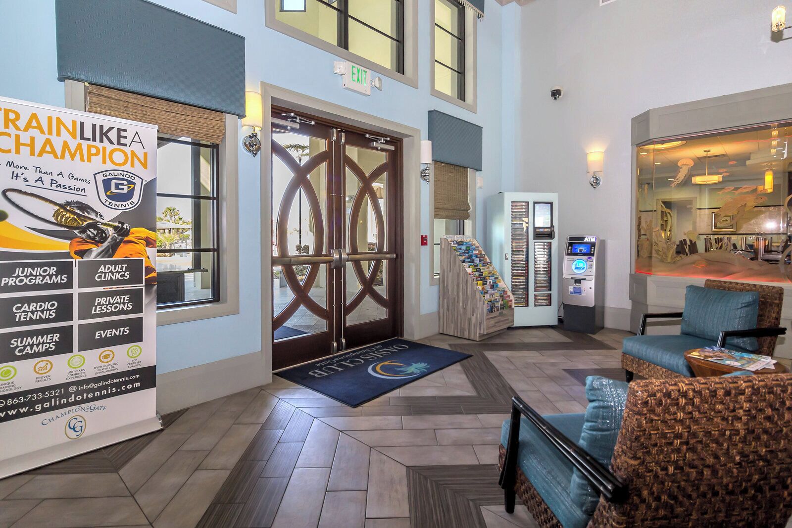 A hotel lobby with a large tennis-themed banner, a reception desk, a staircase, and a seating area with a blue sofa.