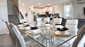 Un área de comedor moderna con una mesa de vidrio, sillas blancas y una cocina al fondo.