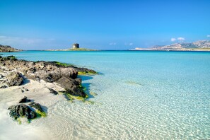 A coastal scene with clear turquoise waters, rocky shoreline, and a distant island with a tower. - Dimora Oleandro 50mt From Beach (Stintino)