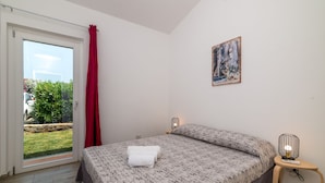 A bedroom with a bed, a nightstand, a painting, and a view of a garden through a sliding door. - Dimora Oleandro 50mt From Beach (Stintino)