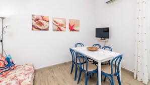 A room with a white table, blue chairs, a TV, and wall art. - Dimora Oleandro 50mt From Beach (Stintino)
