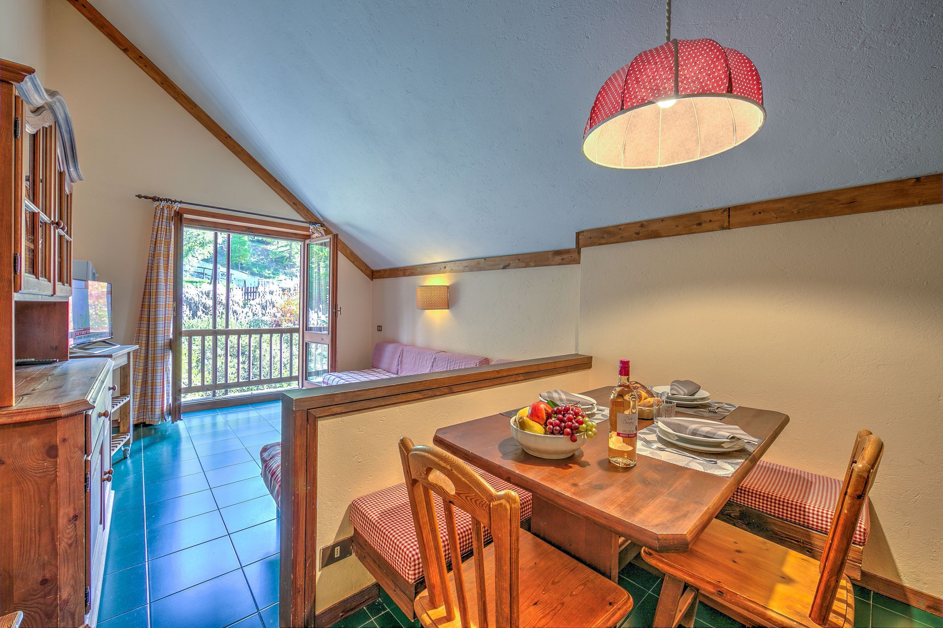 A dining area with a wooden table set for two, a bowl of fruit, and a bottle of wine. There is a window with a view of greenery outside.