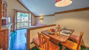 Un coin repas avec une table en bois dressée pour deux personnes, un saladier rempli de fruits et une bouteille de vin. Une fenêtre offre une vue sur la verdure environnante.