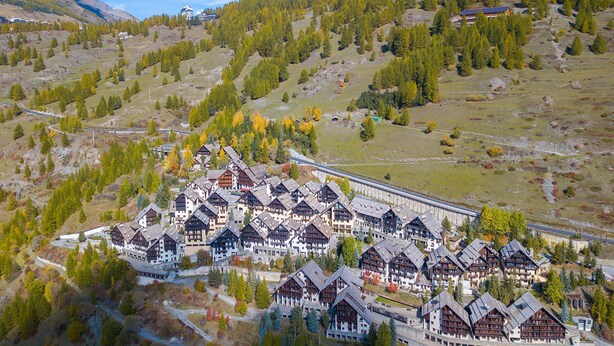 A cluster of traditional chalet-style buildings with a central pedestrian walkway, surrounded by a mountainous landscape with autumn foliage.