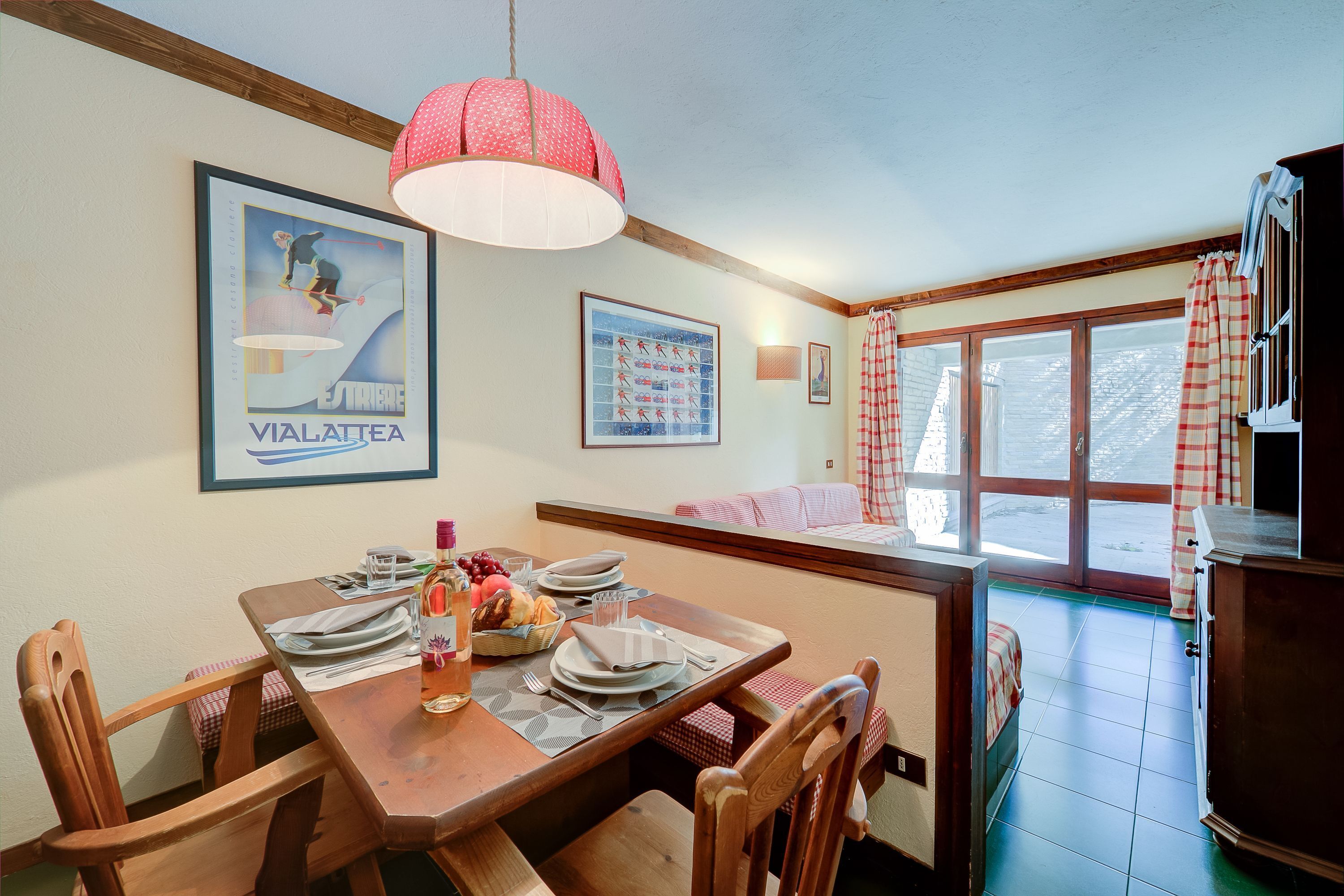 A dining area with a wooden table set for four, a hanging light fixture, and a window with curtains.