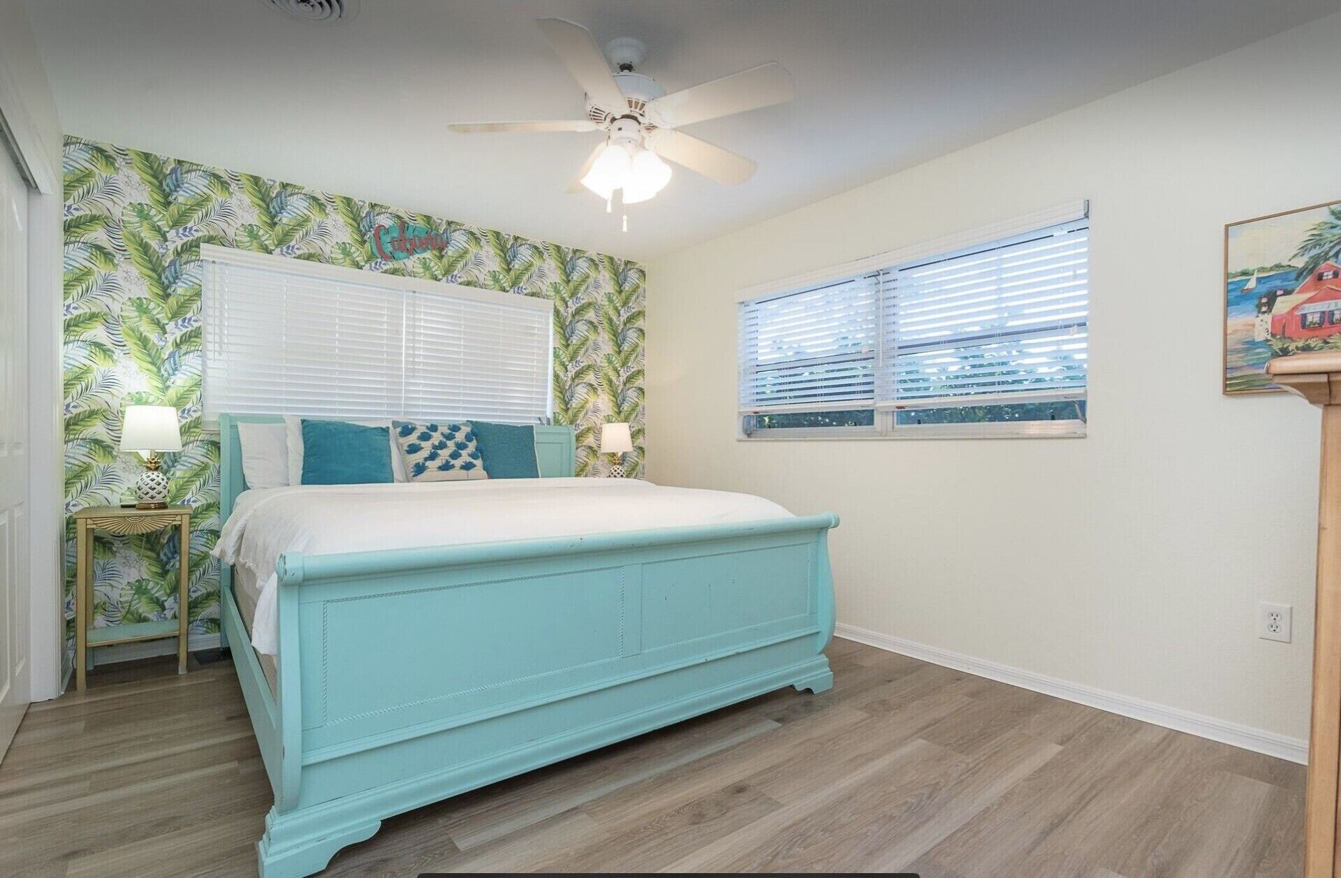 Un dormitorio con cama, ventilador de techo, ventana con persianas y papel tapiz tropical.