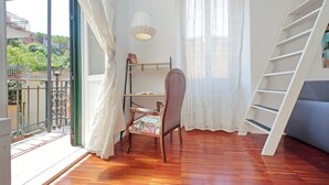Una habitación con piso de madera, una silla, una escalera, una ventana con cortinas y un balcón con barandilla.