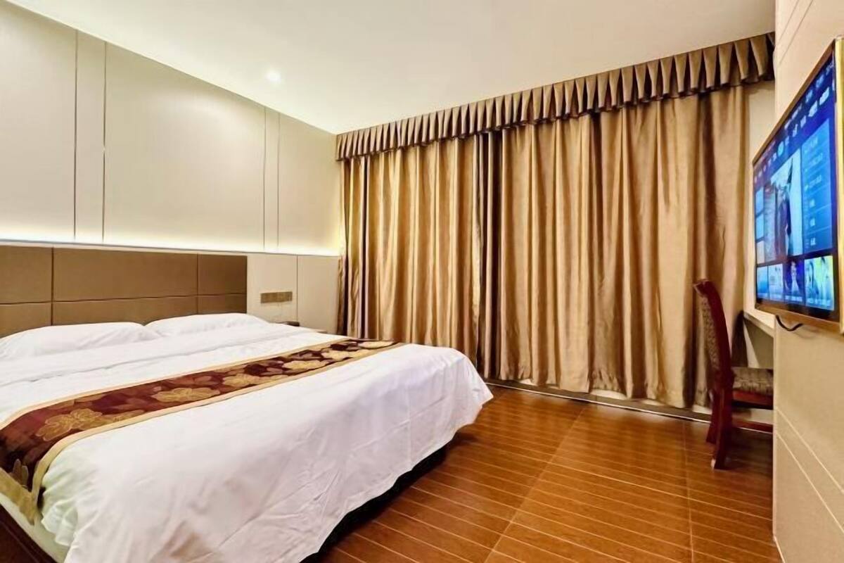 A hotel room with a large bed, a headboard, a TV mounted on the wall, and a window with curtains.