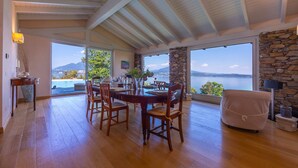 A dining area with a wooden table and chairs, a stone wall, and a view of a body of water. - Paradis Relais Pool View EV Recharge (Belgirate)