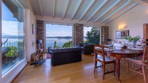 A spacious living area with a large window overlooking a lake, a stone pillar, and a dining table set for four. - Paradis Relais Pool View EV Recharge (Belgirate)