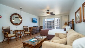 Ein Hotelzimmer mit Bett, Sofa, Esstisch mit Stühlen, Fernseher, Deckenventilator und einem großen Fenster mit Blick auf Gebäude.