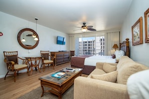 Une chambre d’hôtel comprenant un lit, un canapé, une table à manger avec des chaises, un téléviseur, un ventilateur de plafond et une grande fenêtre donnant sur des immeubles.
