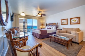 A hotel room with a balcony, a bed, a sofa, a dining table with chairs, and a ceiling fan.