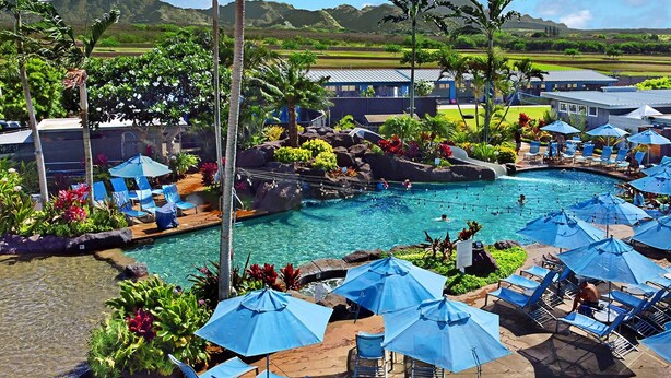 Un espace piscine aménagé dans un complexe hôtelier, avec de nombreux chaises longues et parasols, entouré d’une végétation luxuriante et offrant une vue sur les montagnes lointaines.