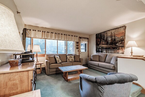A cozy living room with a wooden coffee table, two sofas, a small table with lamps, and a large window with curtains. - One Bedroom Cascades A-24, direct access from outside, sleeps 6 (Newry)