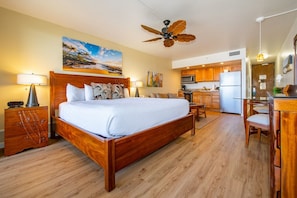 Quarto com cama de madeira, ventilador de teto, uma pintura e área de cozinha com armários e eletrodomésticos de madeira.