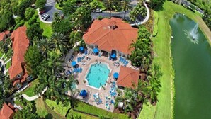 An aerial view of a resort with a central swimming pool surrounded by lounge chairs and umbrellas, a small pond, and a red-roofed building.