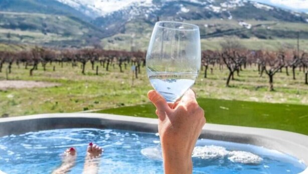 Una mano sosteniendo un vaso de agua sobre un jacuzzi, con un paisaje montañoso al fondo.