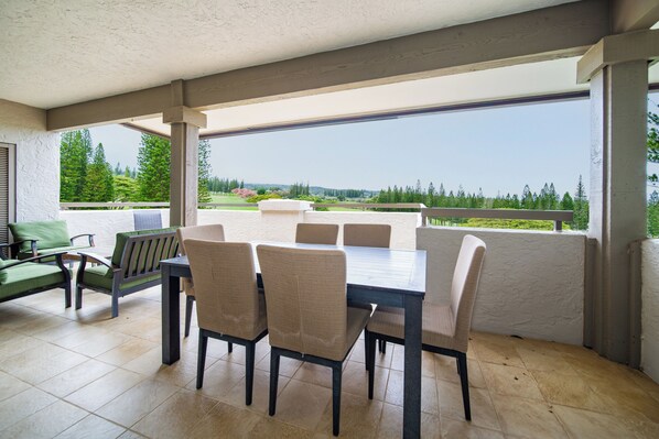 A covered patio with a dining table and chairs, a sofa, and a view of a green landscape.