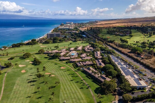 An aerial view of a golf course with a hotel, solar panels, and a coastal backdrop.