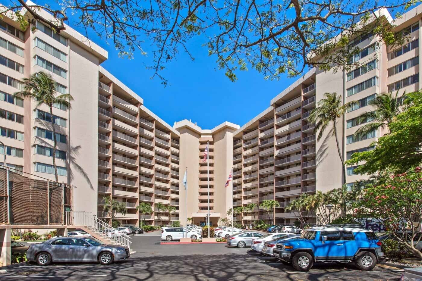 A multi-story hotel with a central courtyard, featuring balconies, a parking area, and palm trees.
