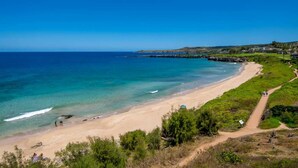 A coastal beach with clear blue waters, a sandy shore, and a path leading to a building.