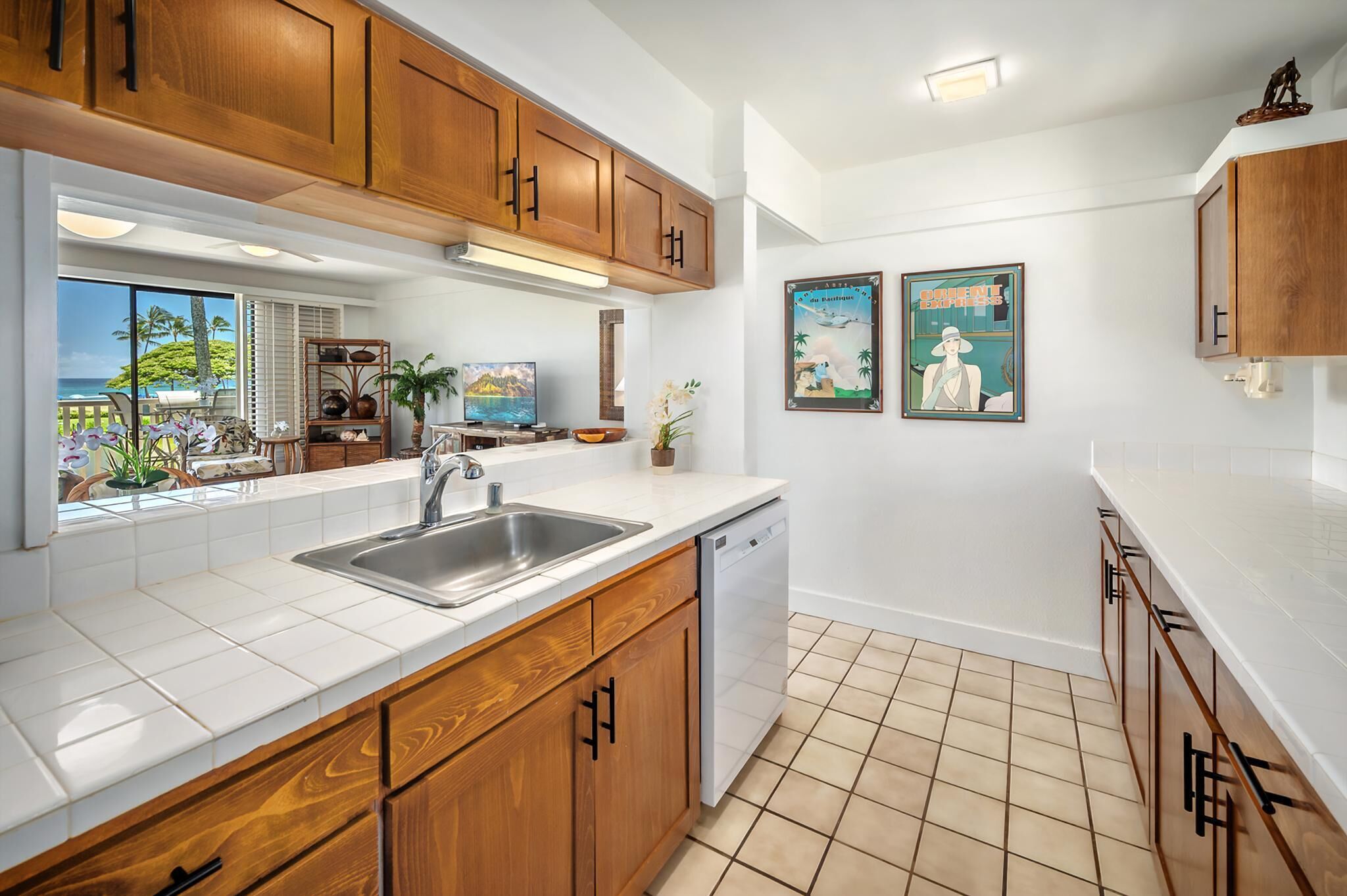 A kitchen with wooden cabinets, a stainless steel sink, and a dishwasher. There are two framed pictures on the wall and a window with a view of palm trees.