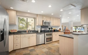 Une cuisine moderne avec des meubles en bois clair, des appareils électroménagers en acier inoxydable et une grande fenêtre équipée de stores.