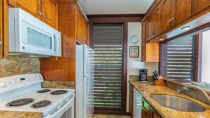 Una cocina con armarios de madera, una estufa blanca, un refrigerador, un microondas, un lavamanos y un ventanal con persianas.