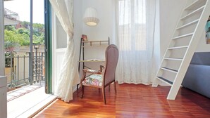 A room with a wooden floor, a chair with a cushion, a bookshelf, a ladder, and a window with curtains. - 4BNB - Angelo Emo - One Bedroom Apartment, Sleeps 5 (Rome)