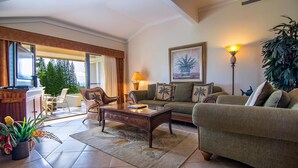 Ein Wohnzimmer mit einer Couch, Sesseln, einem Couchtisch und einer Lampe. Es gibt ein Fenster mit Vorhängen, eine Pflanze und ein Gemälde an der Wand.