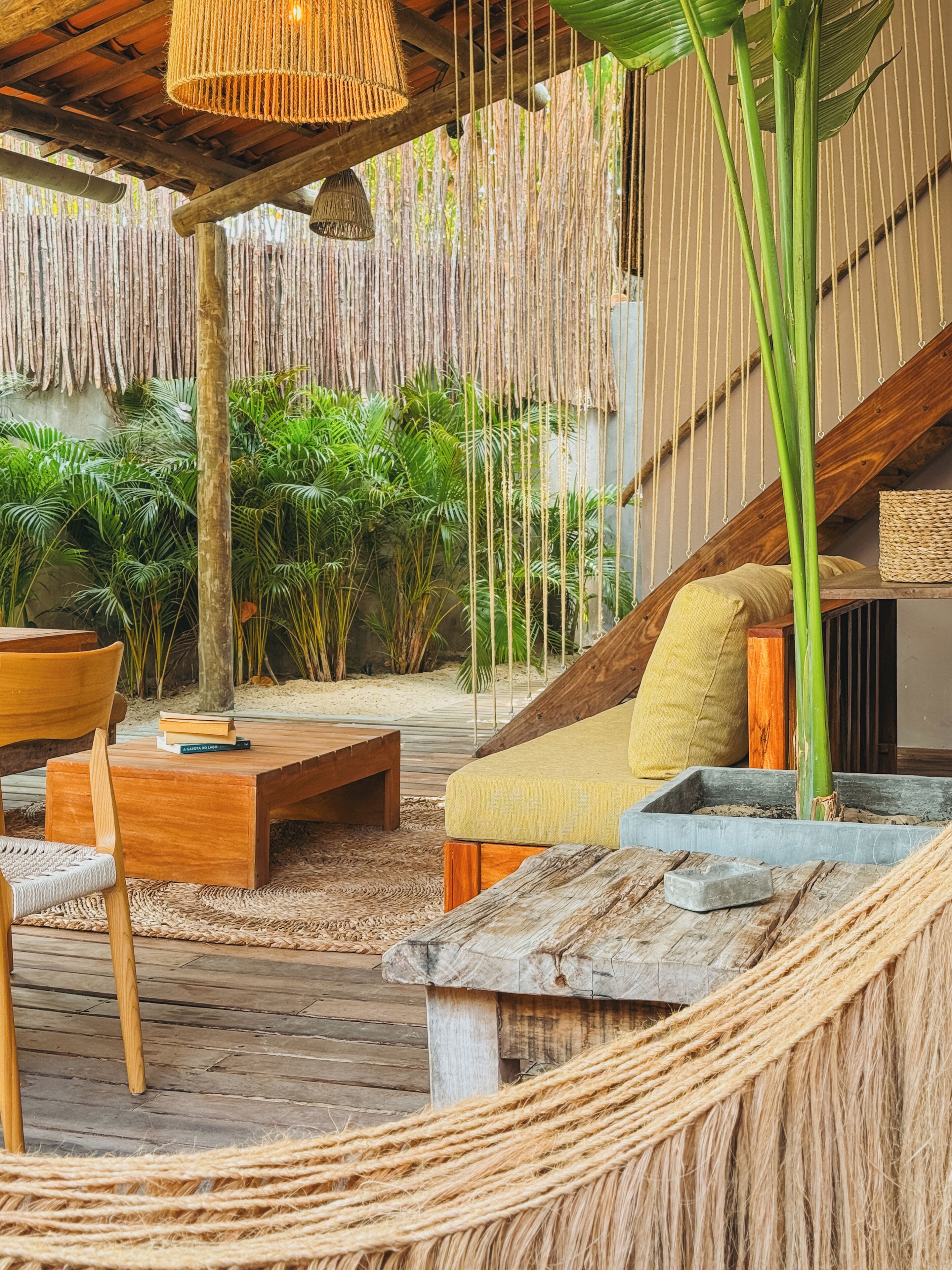 Una terraza de madera con una hamaca tejida, un banco de madera y una zona de estar acolchada con cojines y una cesta tejida.