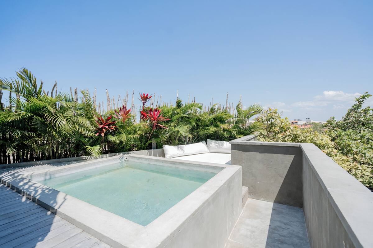 Un espace piscine aménagé sur le toit, avec une piscine rectangulaire entourée d’une végétation luxuriante et de mobilier d’extérieur blanc.