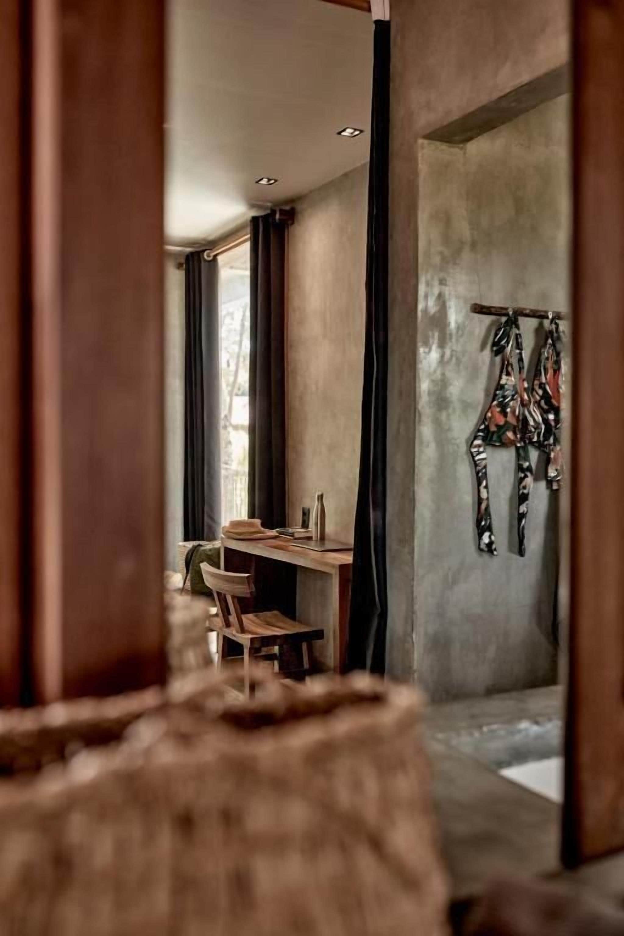 Una stanza con parete in cemento, un tavolo in legno e una sedia. Si vede una finestra con tende e un capo di abbigliamento appeso.
